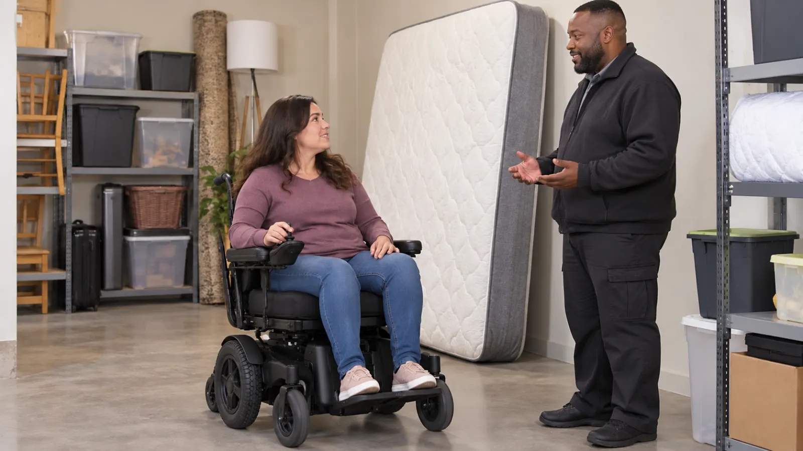 Woman in a power wheelchair speaking with a man beside an upright mattress in a storage room
