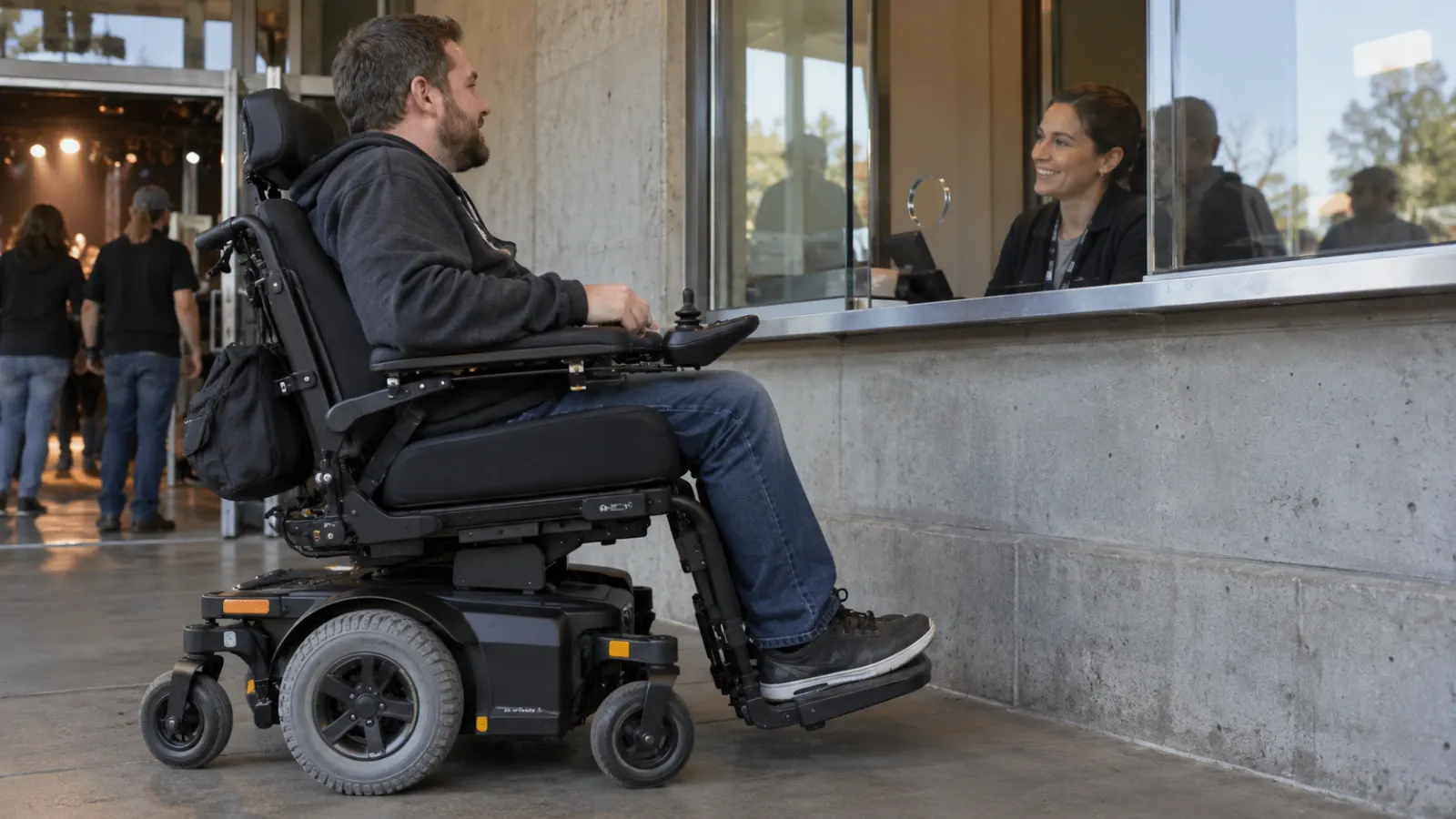A power wheelchair user speaks with a ticket clerk at a venue window.
