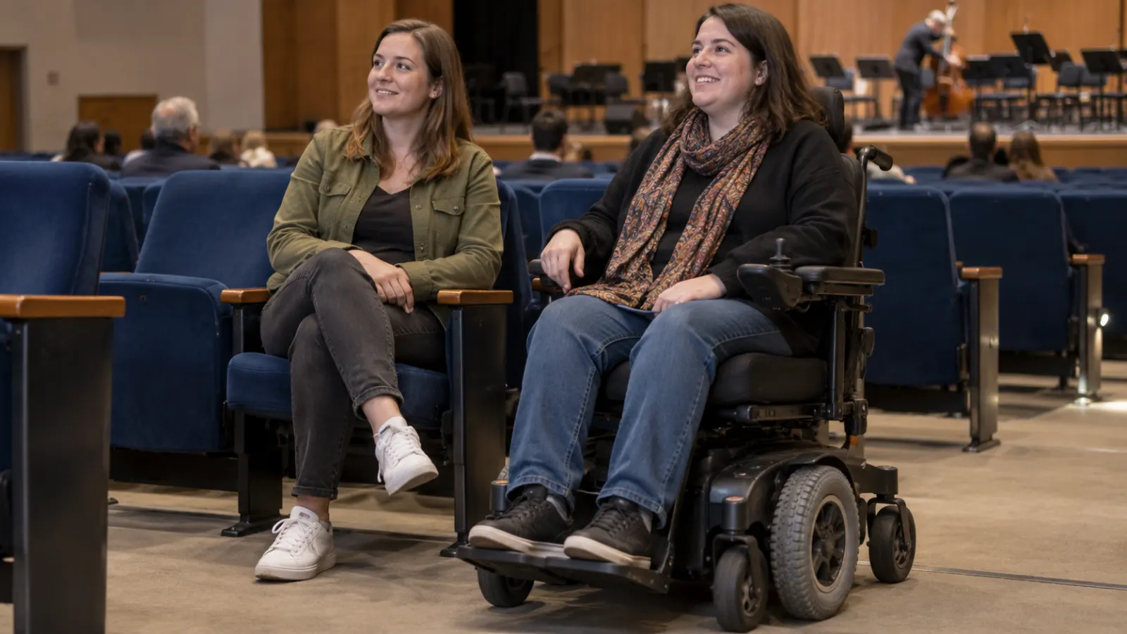 A power wheelchair user sits beside a companion in an auditorium.