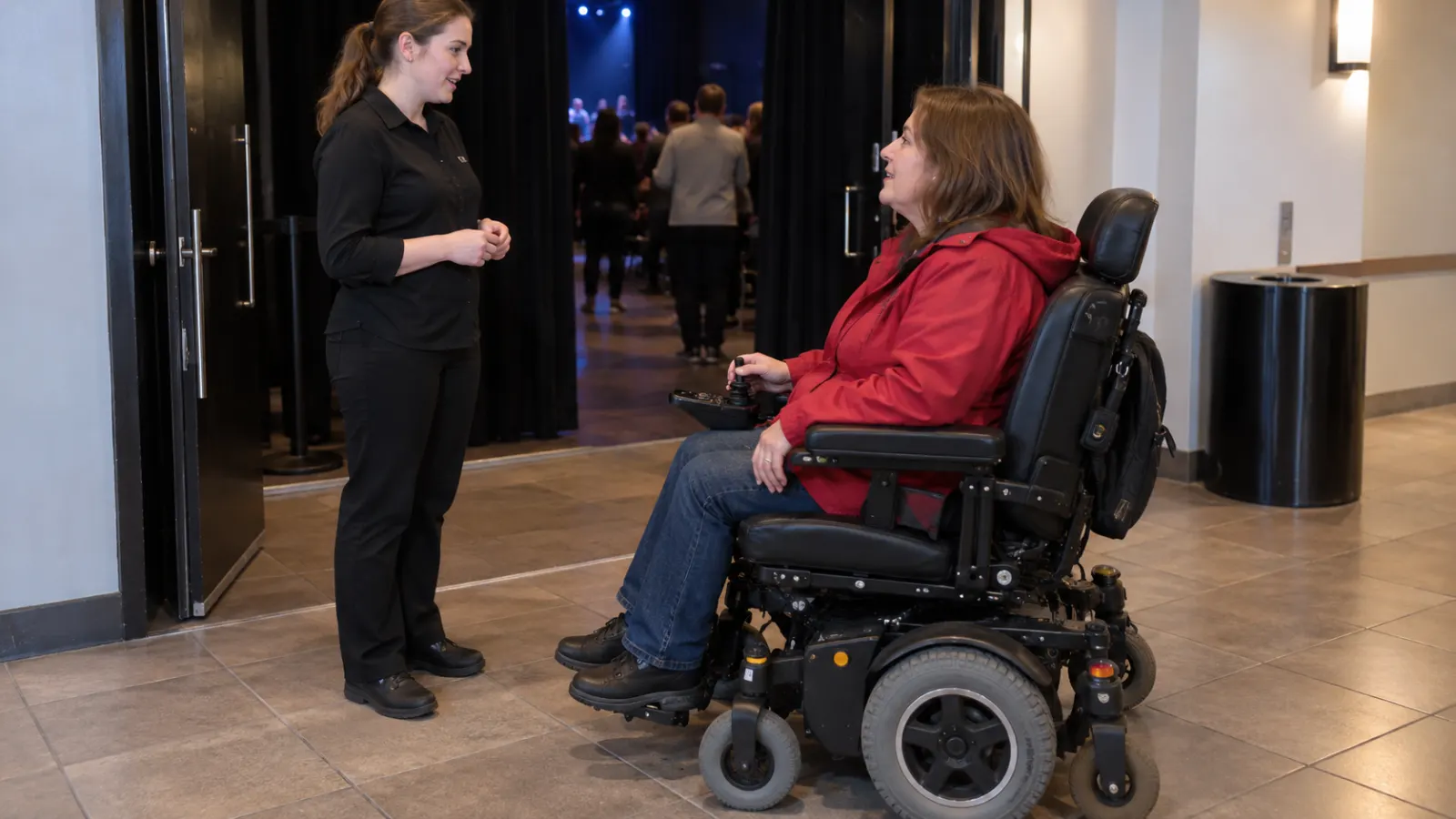 A power wheelchair user talks with an usher near an auditorium door.