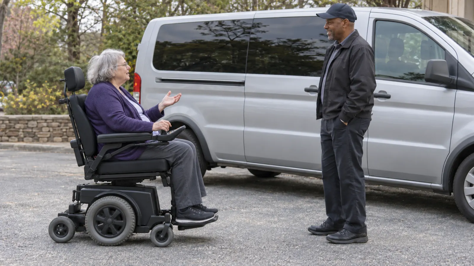 Power wheelchair user speaking with a man beside a parked silver passenger van.