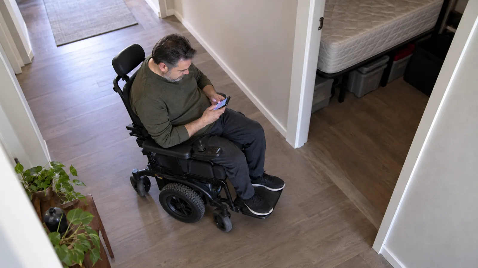 Man in a power wheelchair checking a smartphone in a hallway with a bedroom mattress visible nearby