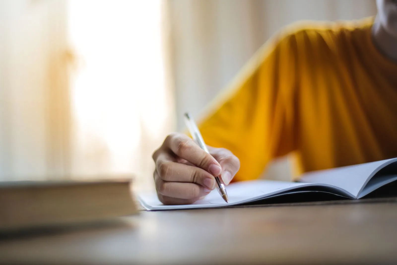 A person's hand holding a silver pen and writing in an open notebook on a wooden table, with a blurred book and yellow shirt visible in the background.