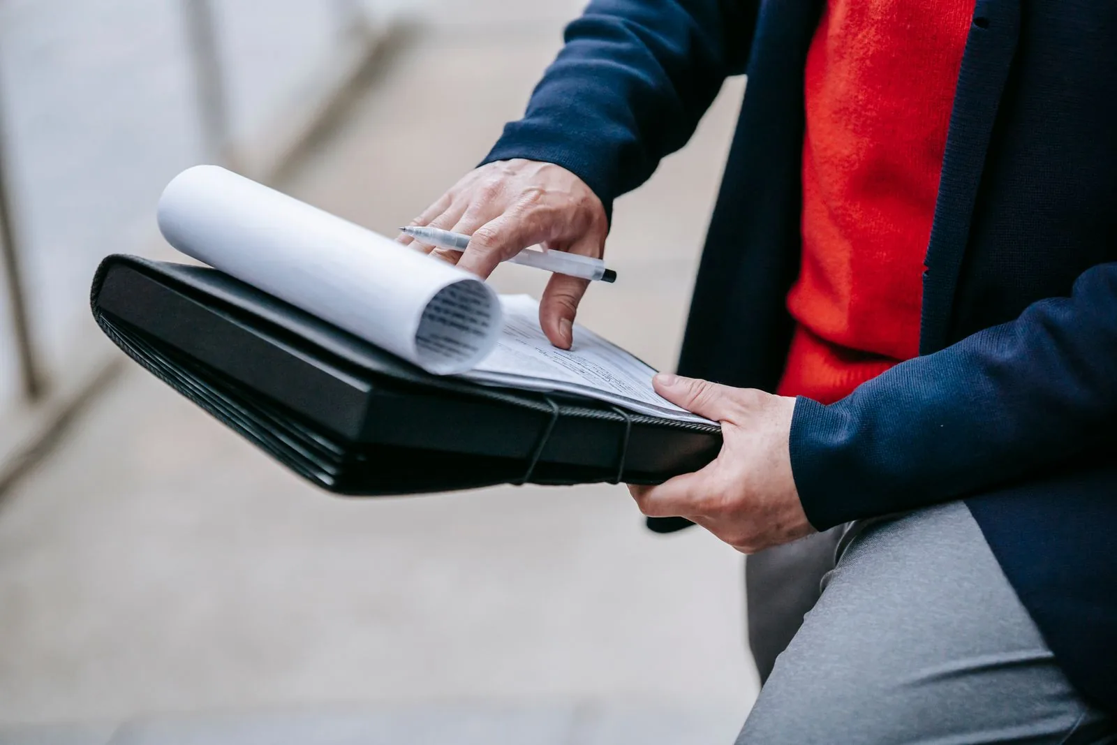 Close-up of hands holding a black portfolio with rolled papers and a pen, wearing a red sweater and navy blazer.