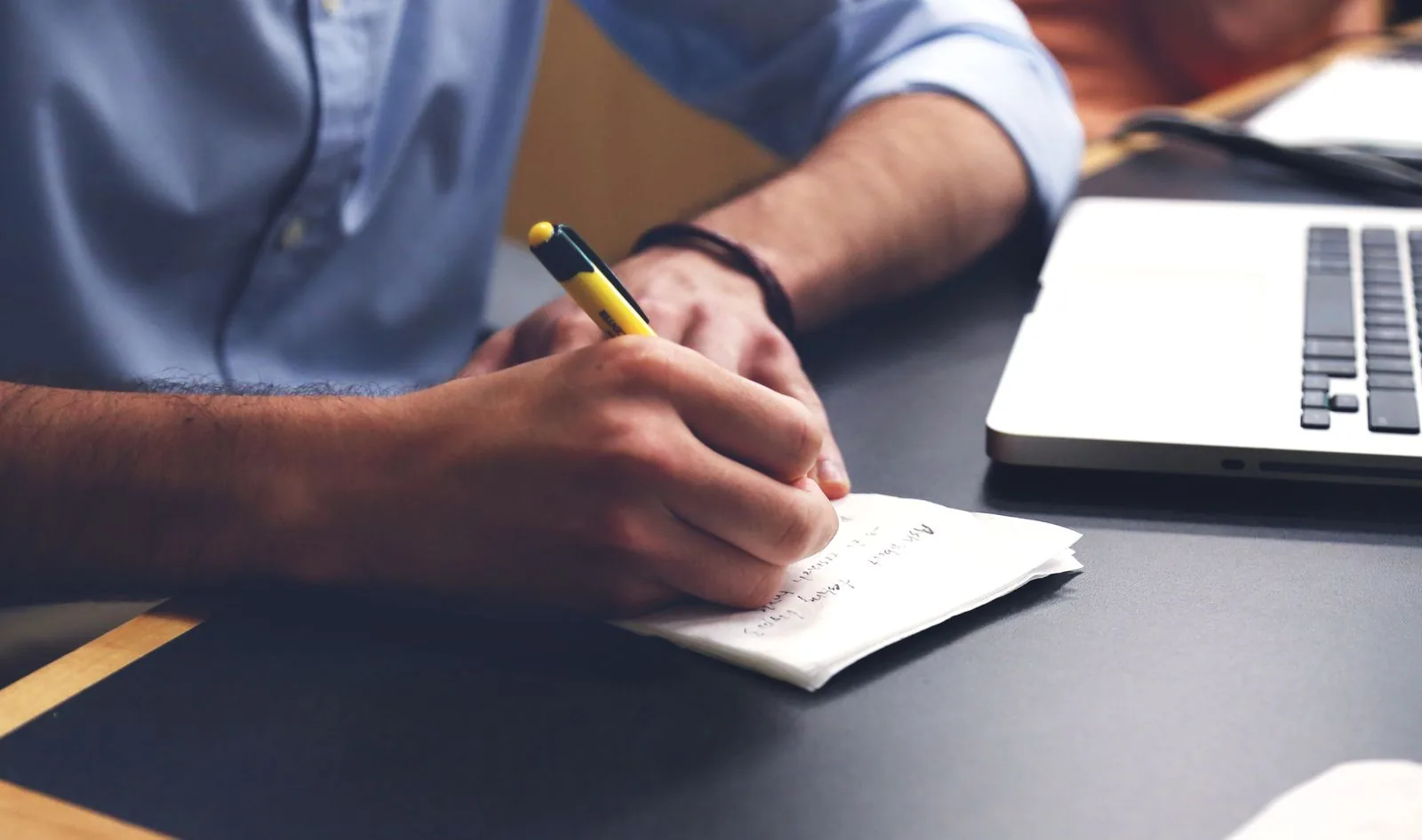 A person's hands writing on a white paper napkin with a yellow pen, a silver laptop visible on the dark desk beside them.
