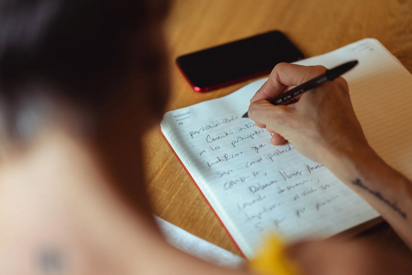 Primer plano de una mano escribiendo notas en cursiva en una agenda diaria abierta con un bolígrafo negro, junto a un teléfono inteligente rojo sobre una mesa de madera.