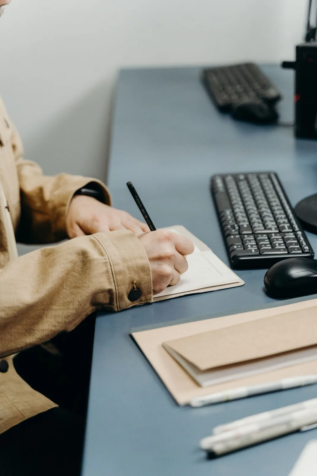 A person's hands writing in a small notebook with a black pencil on a blue desk next to a keyboard and mouse.