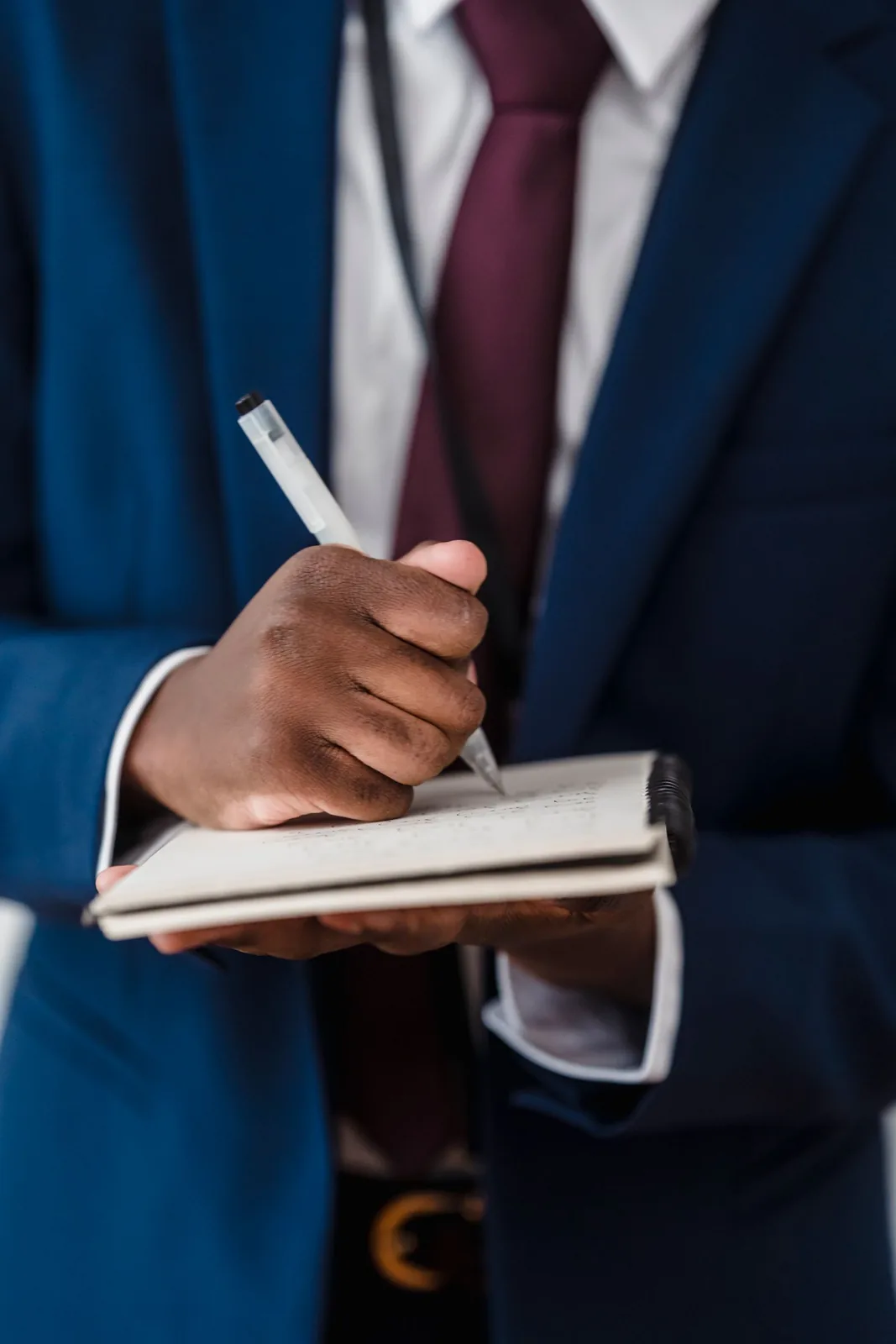 Close-up of a person in a navy blue suit and maroon tie writing on a small notepad with a clear pen.