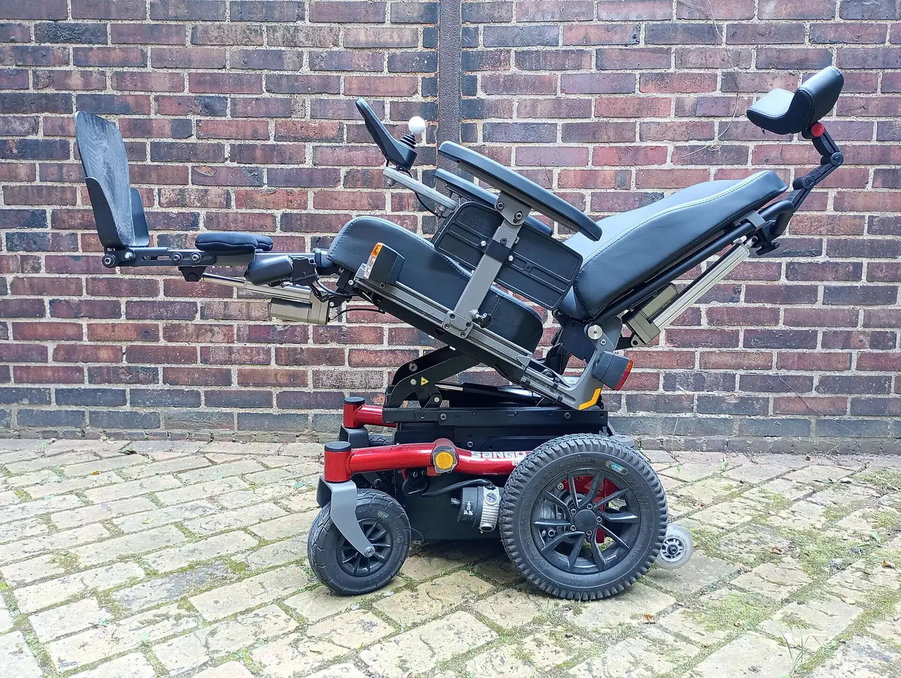 Side view of a red and black electric wheelchair in a reclined position with an extended leg rest, set against a brick wall.