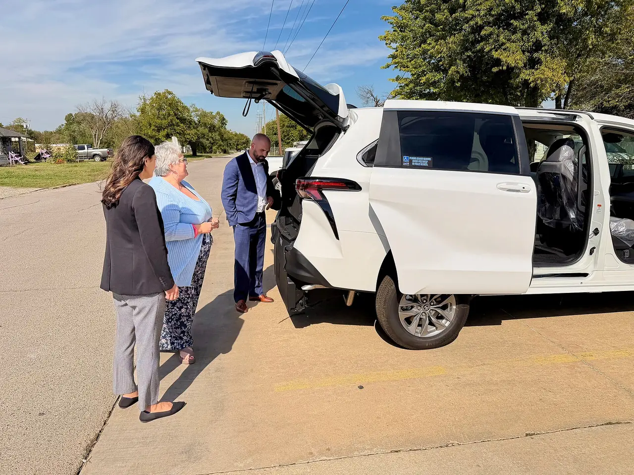 Tres personas están de pie en una carretera pavimentada examinando la parte trasera de un minibús blanco con el maletero y la puerta lateral abiertos, que revelan características de accesibilidad para sillas de ruedas.