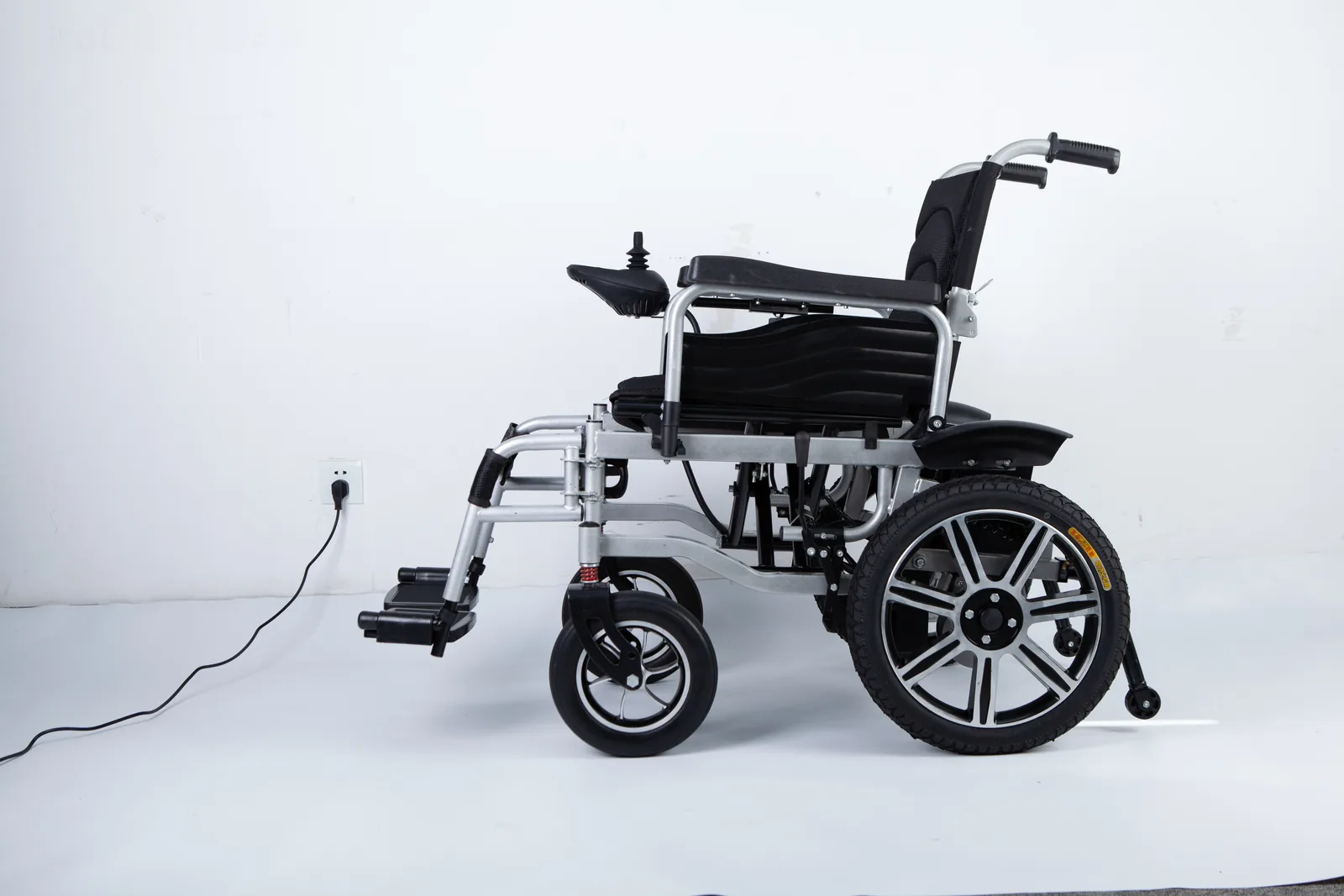 Side view of a silver electric wheelchair with a black seat and a cable running to a wall outlet.