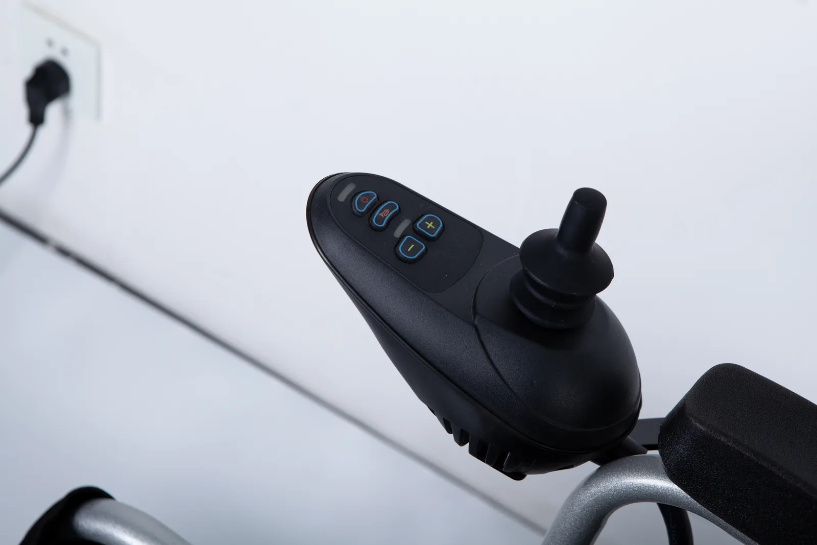 Close-up of a black electric wheelchair joystick controller featuring speed adjustment buttons and a power switch mounted on a silver armrest.