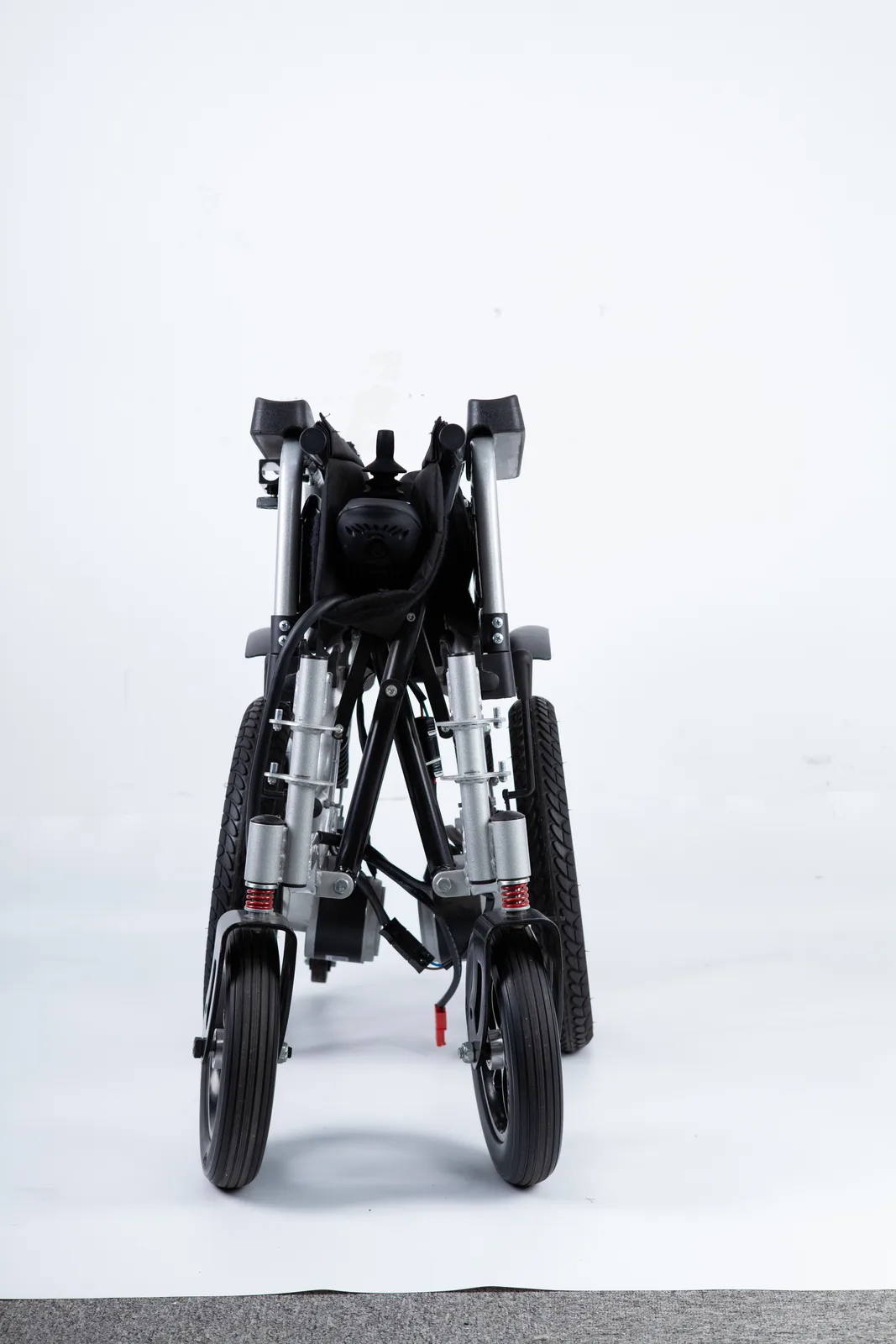 Vue de face d'un fauteuil roulant électrique plié avec de petites roulettes avant, des ressorts de suspension rouges et de grands pneus arrière.