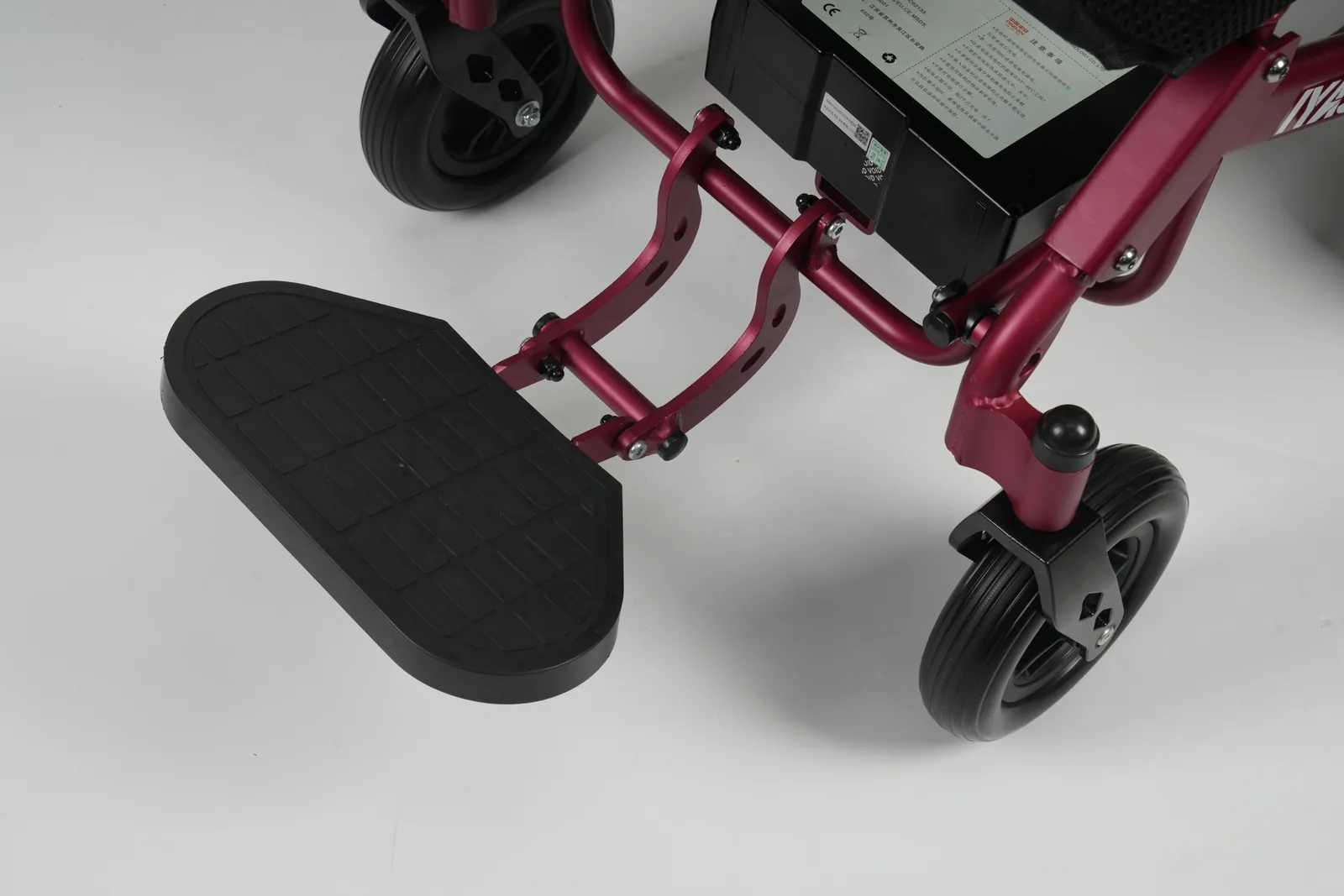 Black textured footplate attached to a maroon metal frame near a small solid rubber caster wheel and battery compartment.