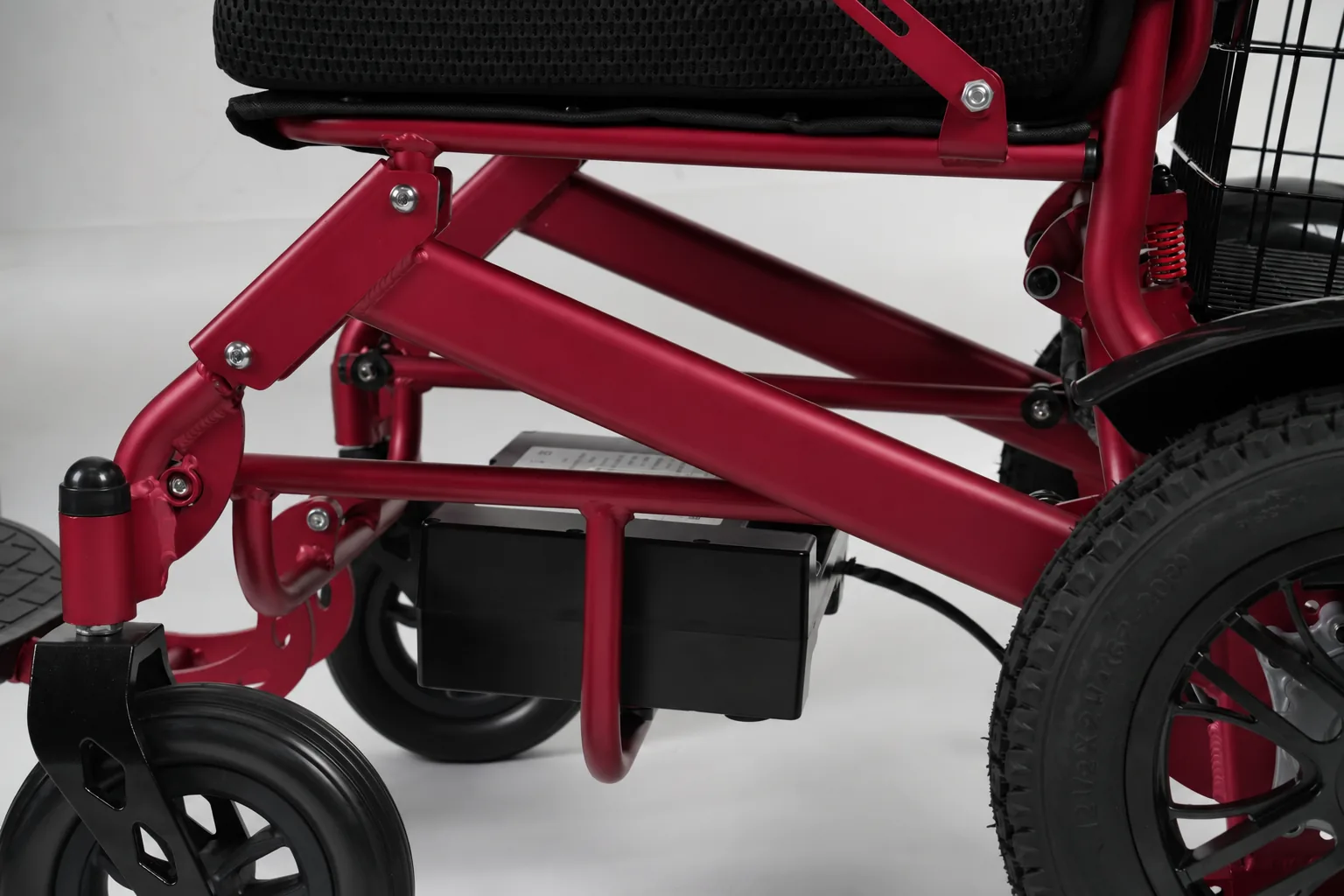 Close-up of a red power wheelchair frame showing the under-seat battery mount, folding mechanism bolts, black mesh seat, and rear pneumatic tire.